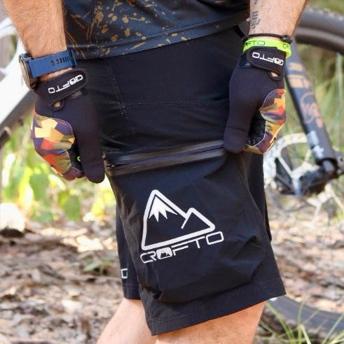 Person wearing a black and brown patterned shirt, black shorts with a logo, and gloves in an outdoor setting.