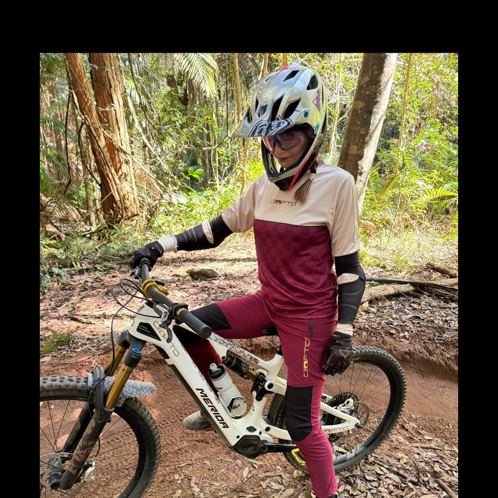 Rider wearing CROFTO Women's Amaroo Lite maroon MTB pants with black panels while seated on a mountain bike on a forest trail.