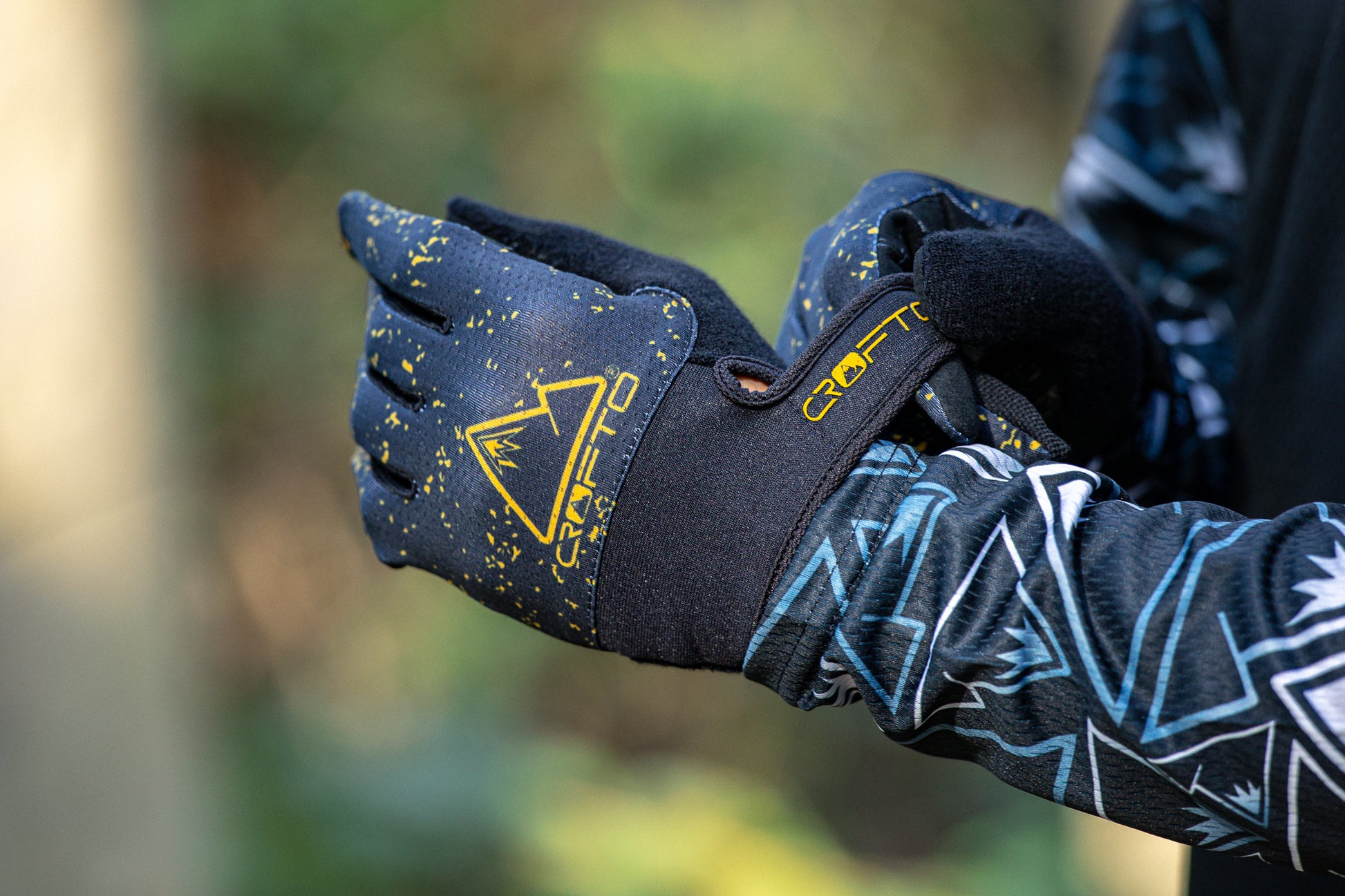 Close-up of a person wearing black and yellow CROFTO Apex Pro mountain biking gloves, with the brand logo and name clearly visible on the back of the glove.