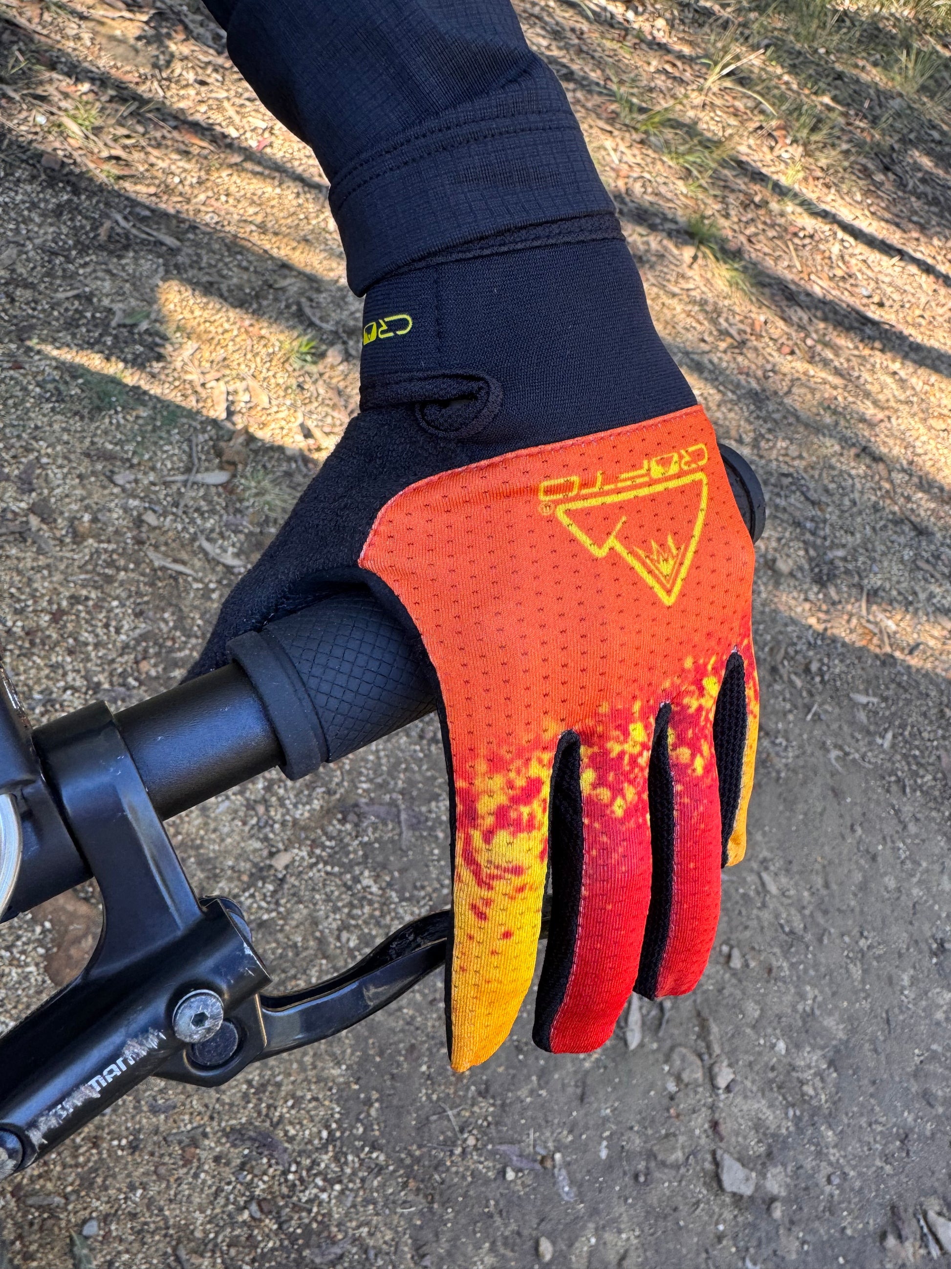 A close-up of a hand wearing a crofto apex pro kids MTB glove with a red, orange, and yellow flame-like pattern, gripping a black bicycle handlebar on an outdoor dirt trail.
