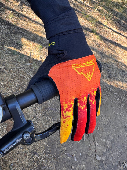 A close-up of a hand wearing a crofto apex pro kids MTB glove with a red, orange, and yellow flame-like pattern, gripping a black bicycle handlebar on an outdoor dirt trail.