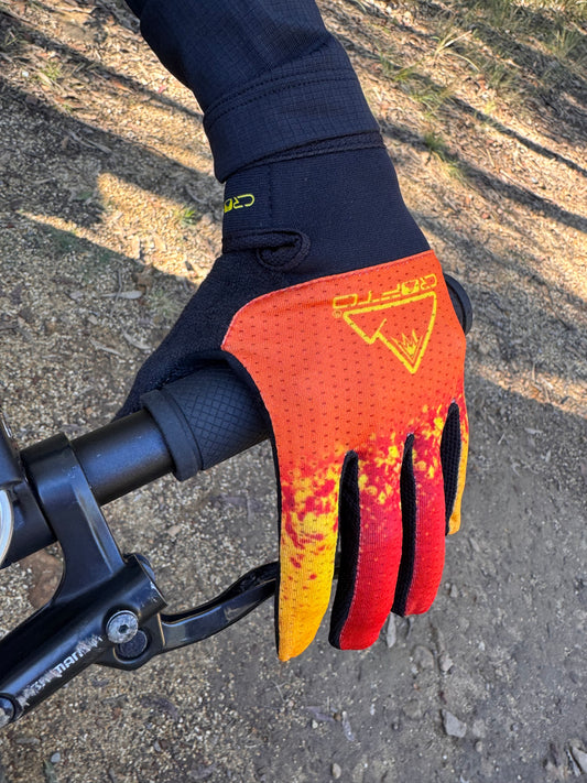 A close-up of a hand wearing a crofto apex pro kids MTB glove with a red, orange, and yellow flame-like pattern, gripping a black bicycle handlebar on an outdoor dirt trail.