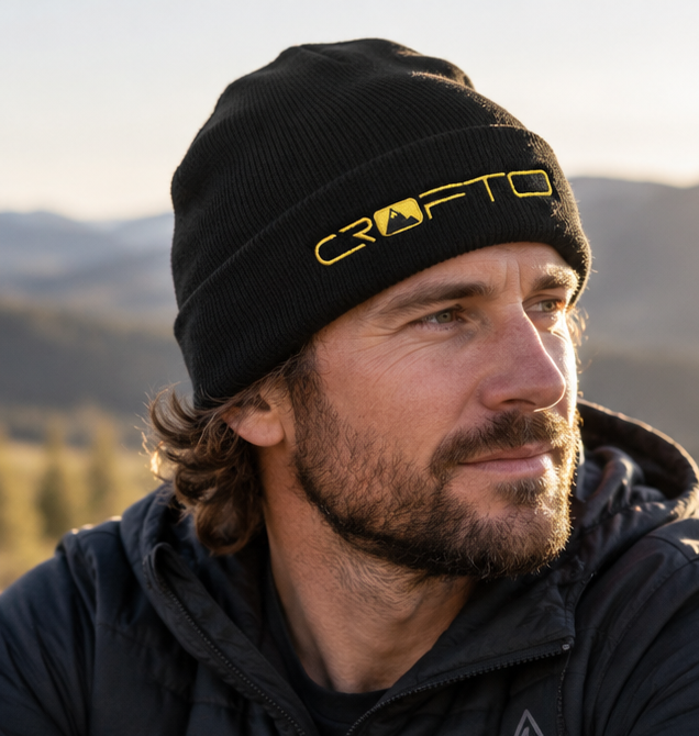 Male mountain biker wearing a black CROFTO knitted beanie with gold embroidered logo in an outdoor mountain setting.