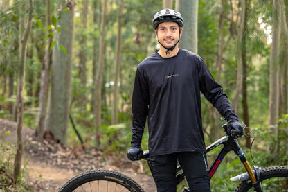 Rider wearing the CROFTO Enduro long sleeve mountain bike jersey in black while standing beside a mountain bike on a forest trail.