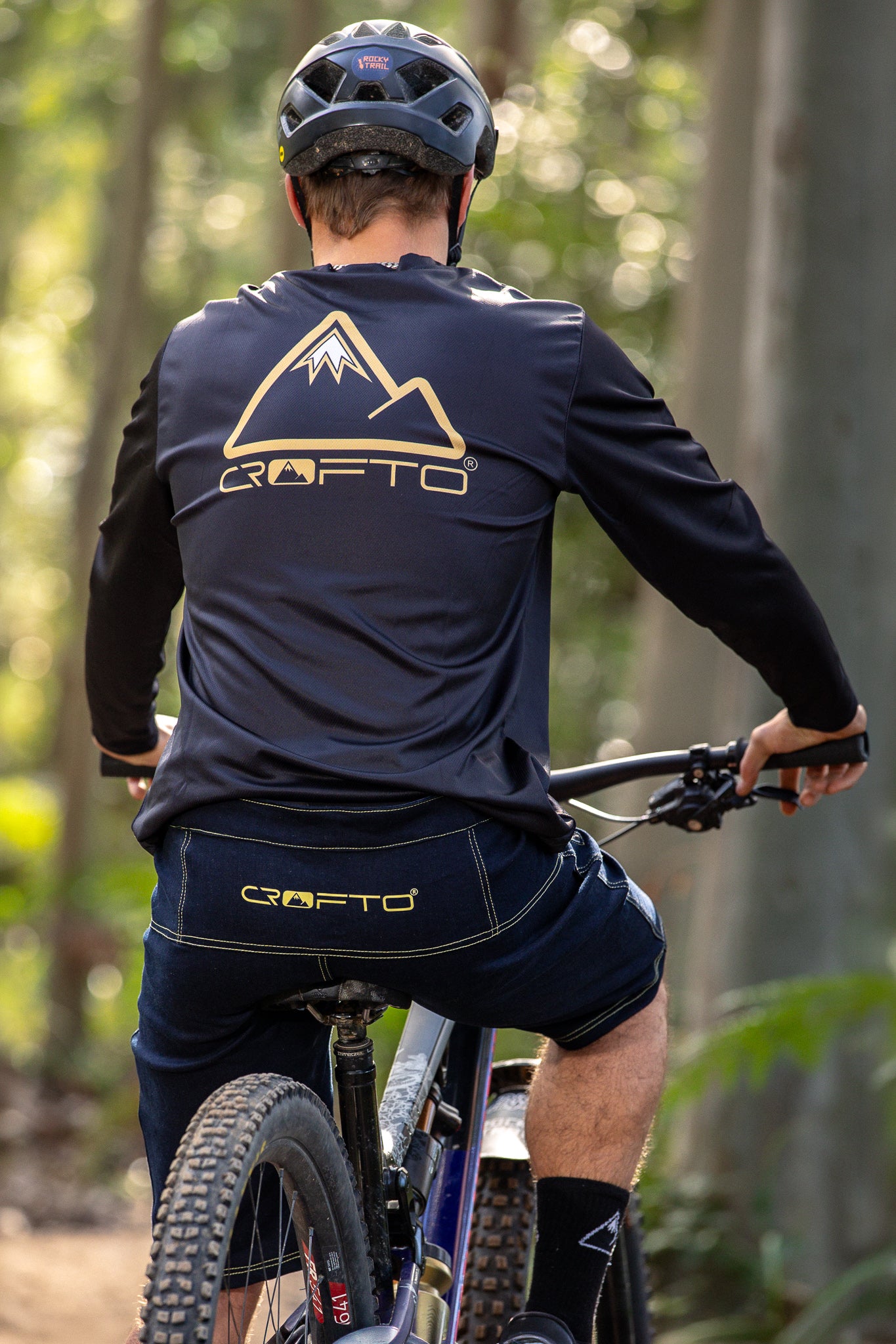 A rear view of a person wearing a CROFTO Enduro MTB Jersey and CROFTO Denim MTB Jorts, Sitting on a mountain bike in the  forest 