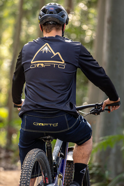 A rear view of a person wearing a CROFTO Enduro MTB Jersey and CROFTO Denim MTB Jorts, Sitting on a mountain bike in the  forest 