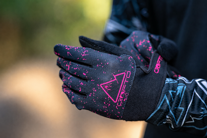 Close-up of black CROFTO mountain bike gloves featuring a pink splatter design and pink CROFTO logo, worn by a rider on a forest trail.