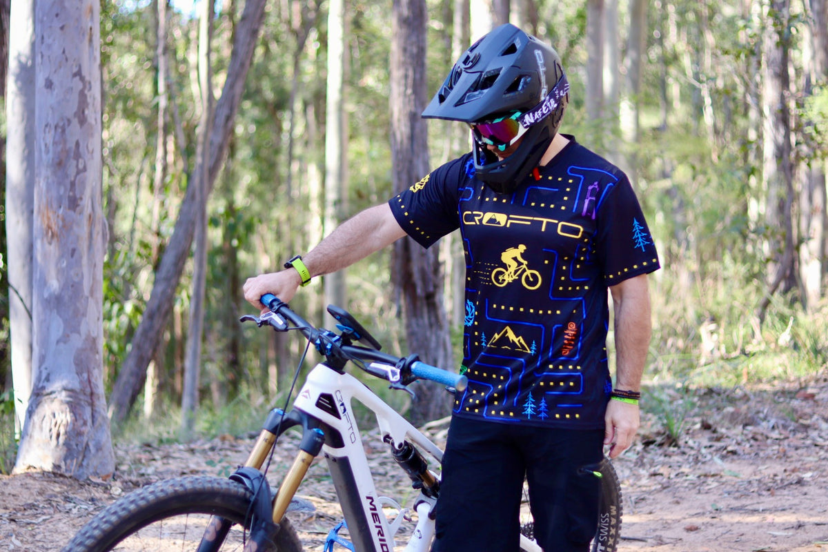Rider wearing the CROFTO Trail Maze mountain bike jersey featuring a retro gaming inspired maze design while standing beside a mountain bike in a forest trail setting.