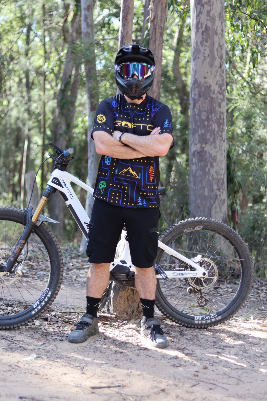 A mountain biker wearing a black helmet, black shorts, and a CROFTO Trail Maze MTB jersey with blue and yellow details stands in a wooded outdoor setting with a white mountain bike behind them.