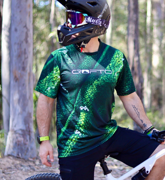 Rider wearing the CROFTO Viper mountain bike jersey featuring a green scale pattern design while trail riding in a forest on a full-suspension mountain bike.