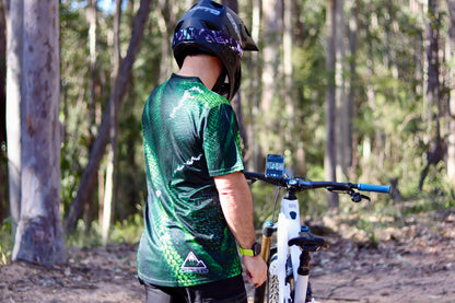 Rider wearing the CROFTO Viper mountain bike jersey featuring a green scale pattern design while trail riding in a forest on a full-suspension mountain bike.