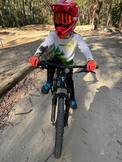 Young rider wearing a CROFTO mountain bike jersey, full-face helmet and gloves with a mountain bike on a forest trail, showcasing youth MTB apparel and trail riding gear.