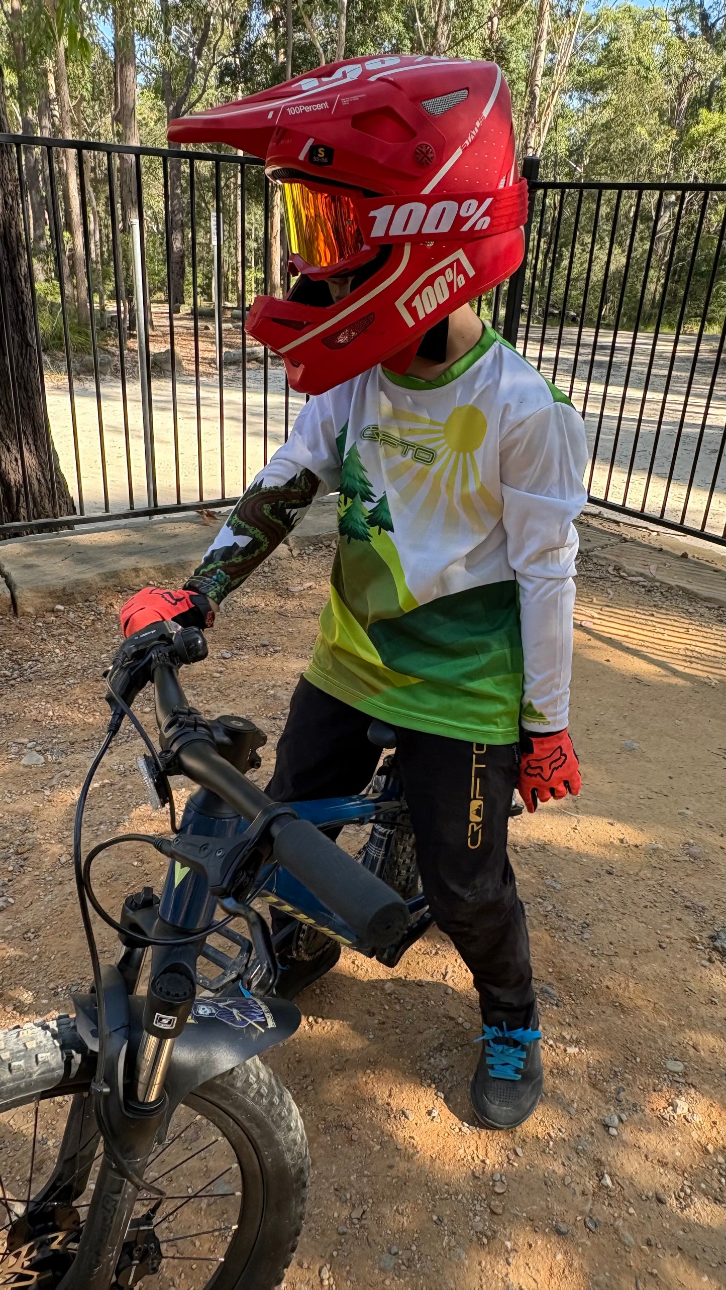 Young rider wearing a CROFTO MTB jersey and full-face helmet with a mountain bike on a dirt trail, showcasing youth mountain biking apparel and trail riding gear.