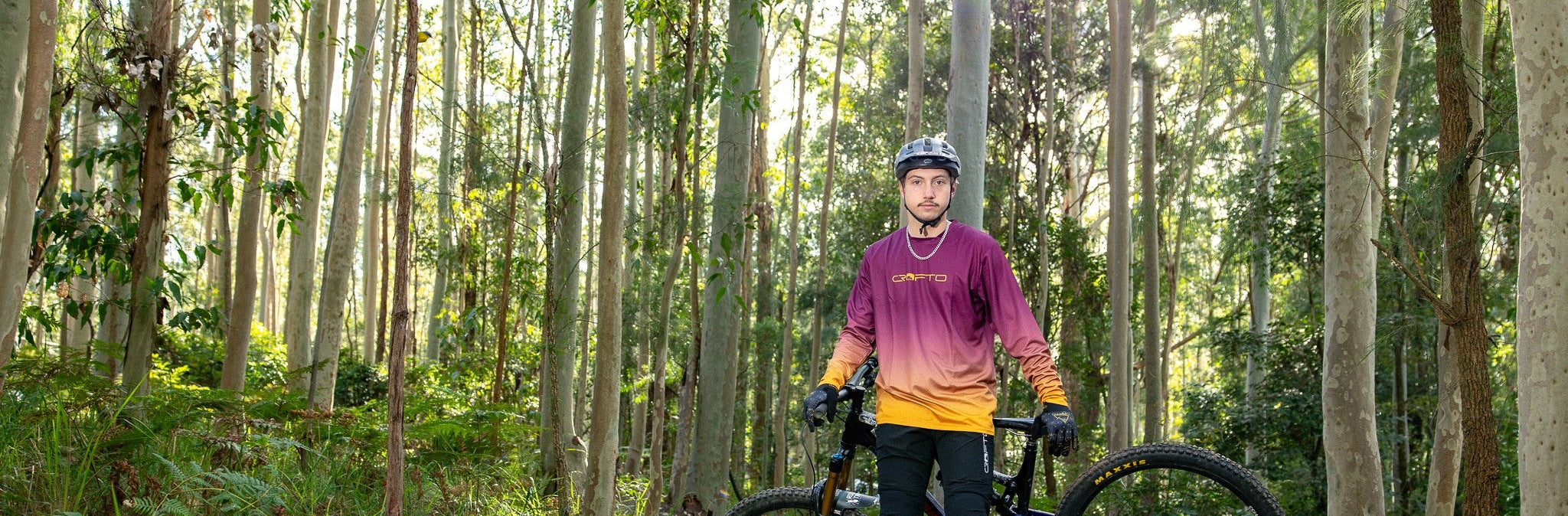 landscape photo of crofto team rider wearing black amaroo pants and crofto fade jersey leaning on his bike in a forest setting