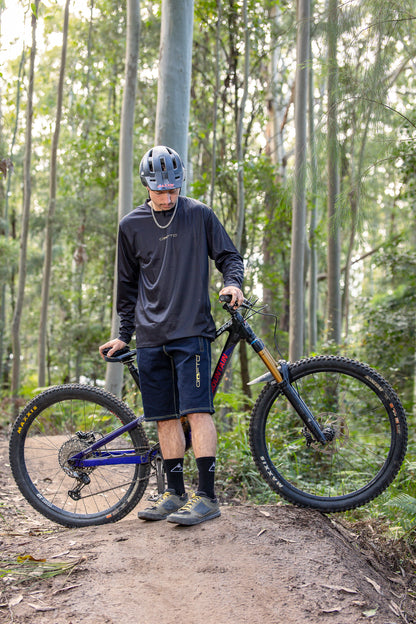 A person wearing a CROFTO Enduro MTB Jersey and CROFTO Denim MTB Jorts, standing next to a mountain bike in a forest setting at Ourimbah