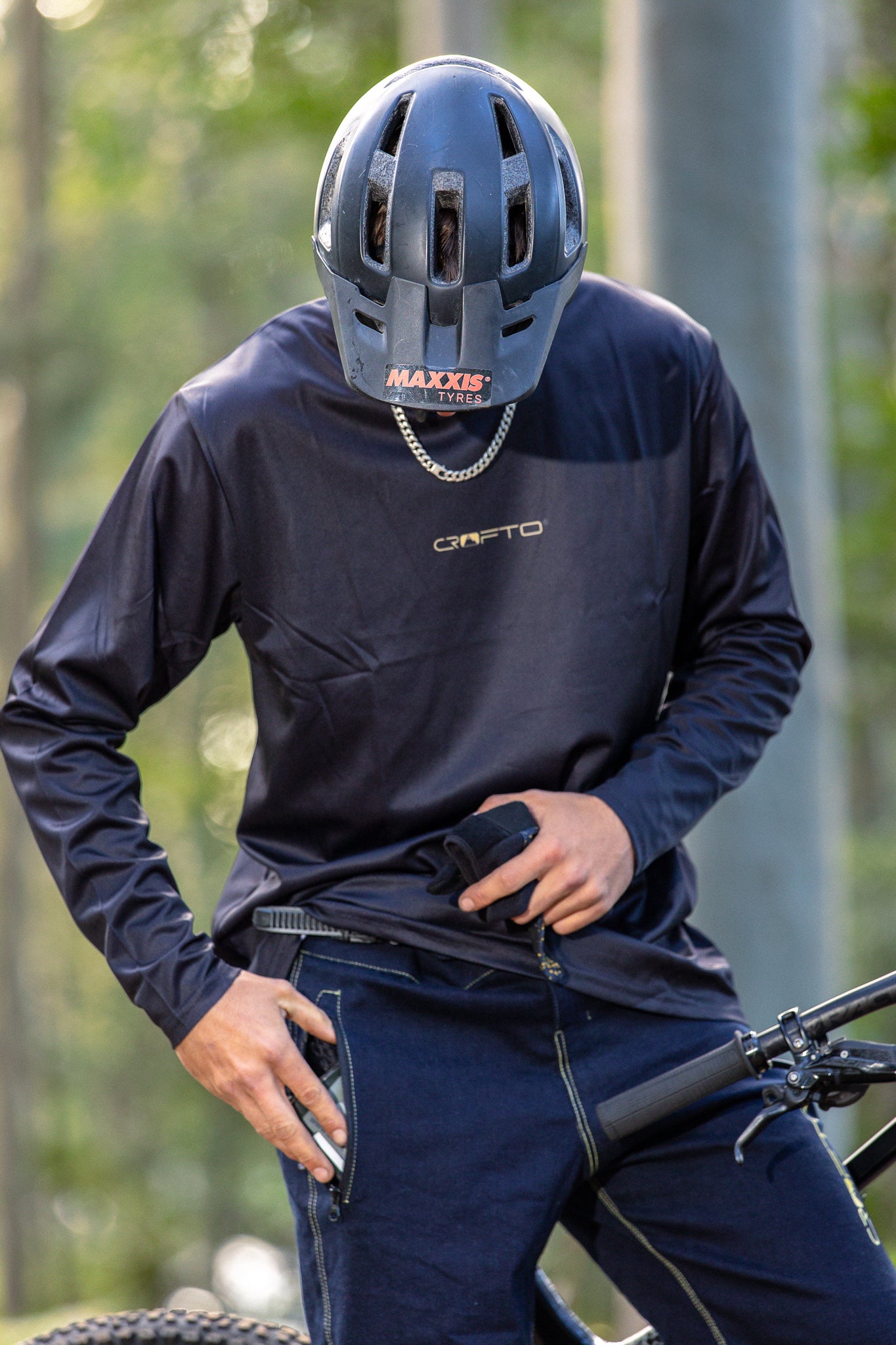 A person wearing a black Maxxis mountain bike helmet, a black long-sleeve CROFTO jersey with a silver chain necklace, and dark blue CROFTO MTB Jorts, standing next to a bicycle in an outdoor setting with blurred trees in the background. 