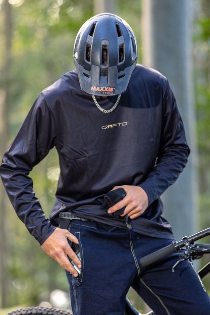 A person wearing a black Maxxis mountain bike helmet, a black long-sleeve CROFTO jersey with a silver chain necklace, and dark blue CROFTO MTB Jorts, standing next to a bicycle in an outdoor setting with blurred trees in the background. 