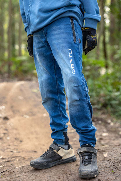 A person wearing blue denim crofto pants with a vertical white CROFTO logo on the left leg, standing on a dirt trail in a forest. The person is also wearing a blue hoodie, black gloves, and black cycling shoes.