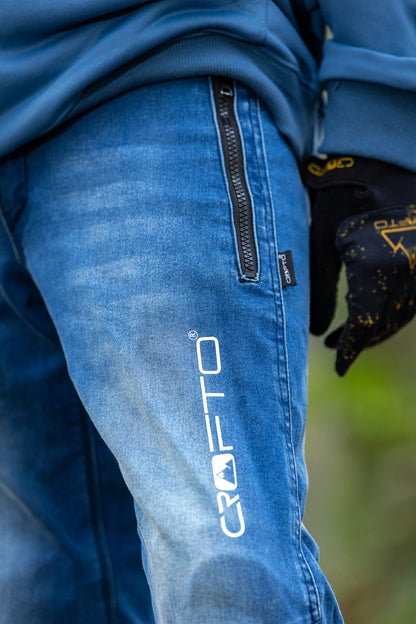 Close-up of a person wearing blue Crofto MTB jeans with a vertical white logo on the thigh, a side zipper pocket, and a black work glove.