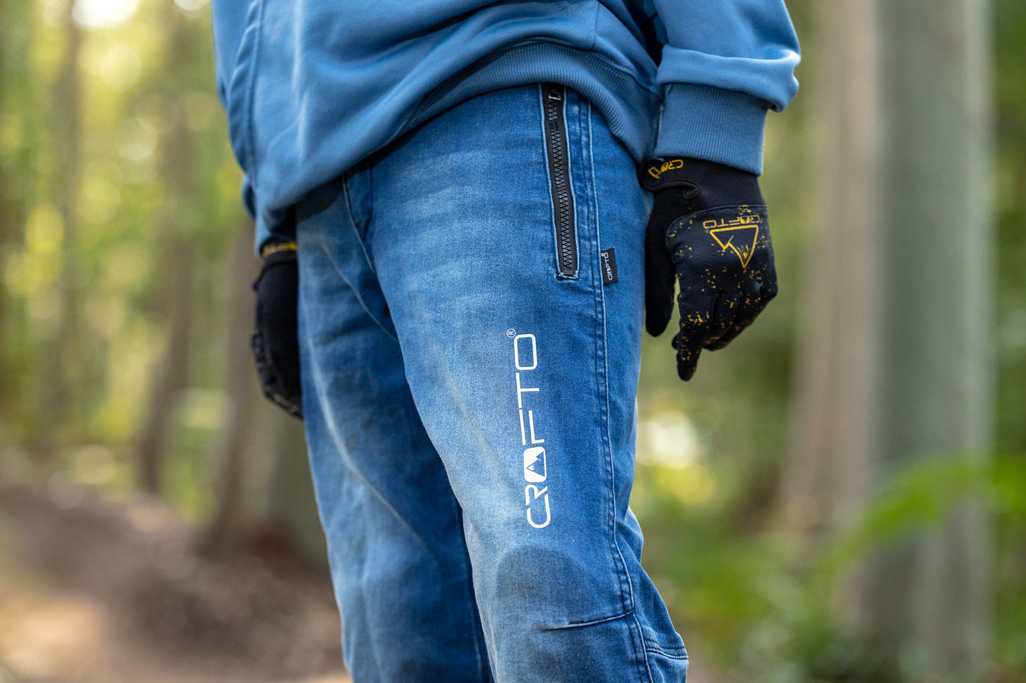 A close-up of a person wearing blue crofto jeans and black gloves in a blurred forest setting. The brand name ‘CROFTO' is printed vertically in white on the left thigh.