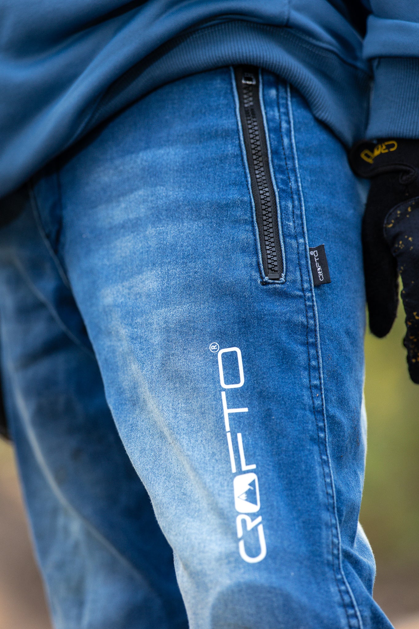 Close-up of a person wearing blue Crofto pants with a vertical white logo on the thigh, a black zipper pocket, and a black glove partially visible.