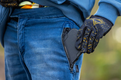 A close-up of a person wearing blue CROFTO MTB Rider jeans and a blue crofto trail hoodie, with a black smartphone tucked into a zippered pocket of the jeans. The person is wearing a black and yellow crofto apex pro glove on the hand.