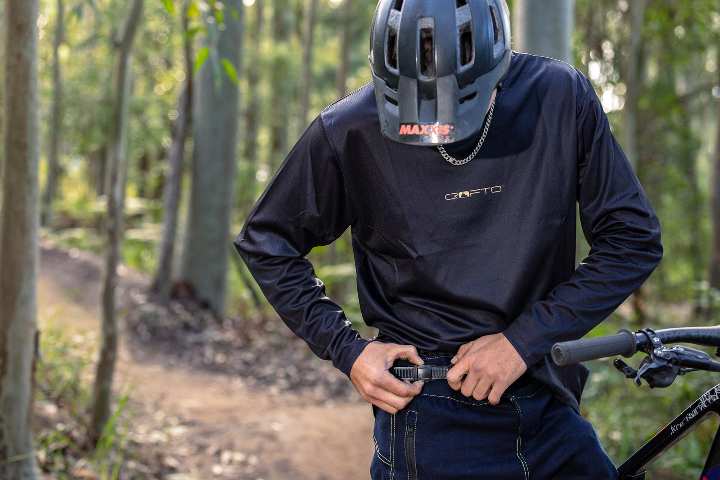 A person wearing a black long-sleeve CROFTO Enduro jersey and CROFTO Denim Jorts stands on a forest trail, adjusting their pants next to a bicycle.