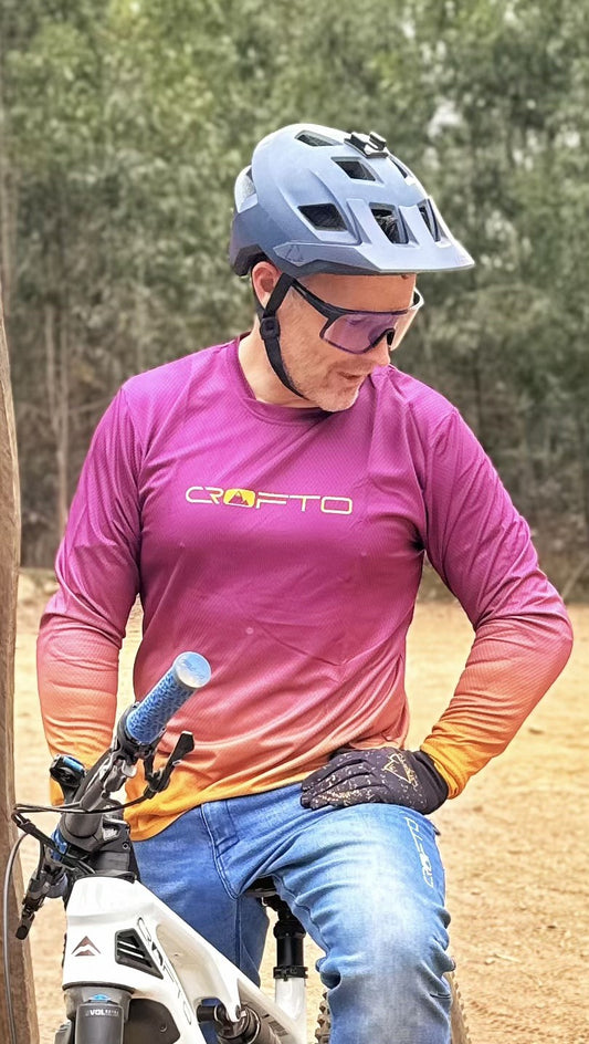 Person wearing a purple shirt with a brand logo, sitting on a bike outdoors.