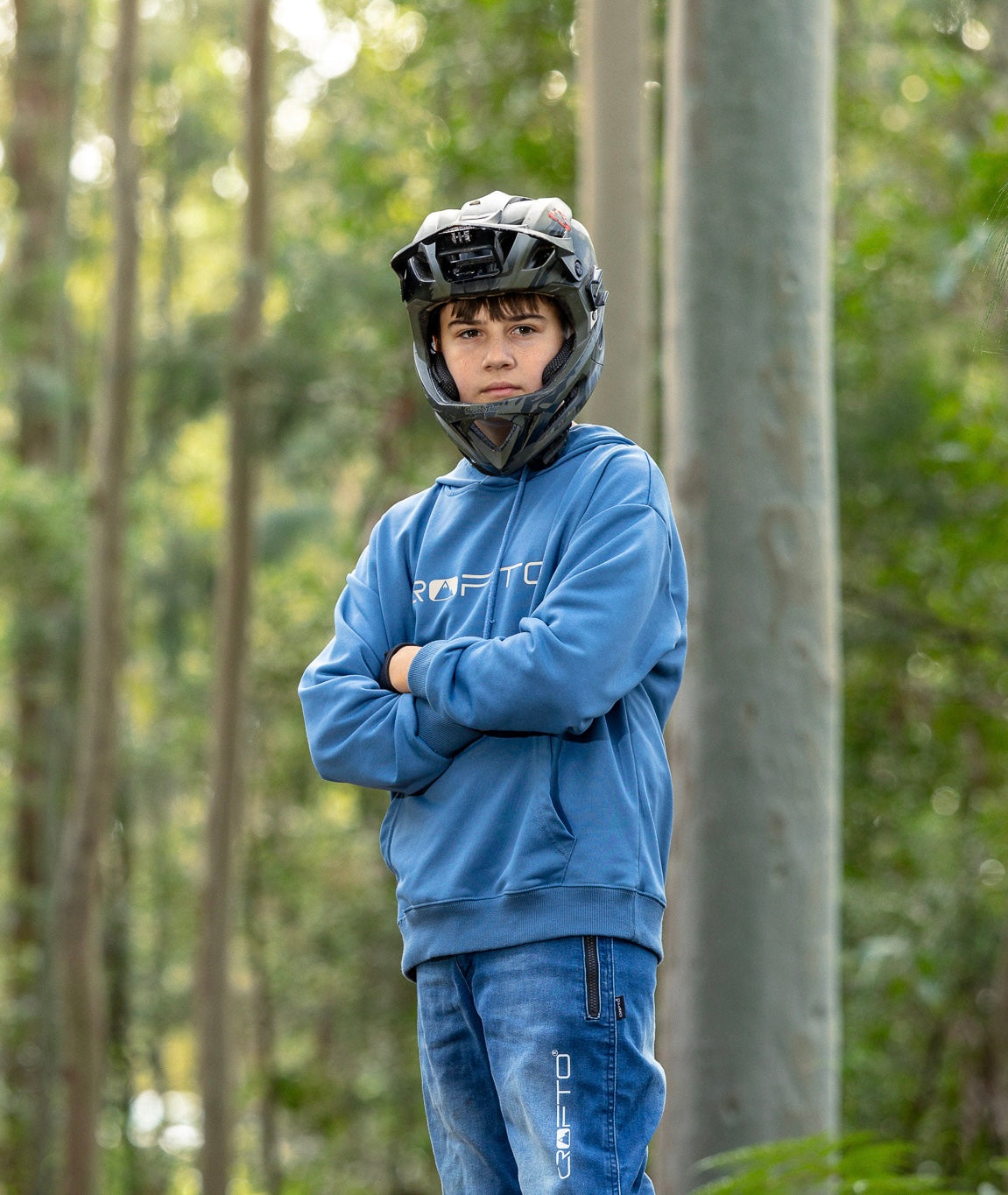 A young boy wearing a blue CROFTO hoodie and matching blue riding jeans stands in a forest, wearing a black full-face helmet with a mounted camera.