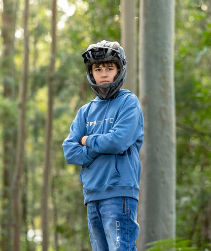 A young boy wearing a blue CROFTO hoodie and matching blue riding jeans stands in a forest, wearing a black full-face helmet with a mounted camera.