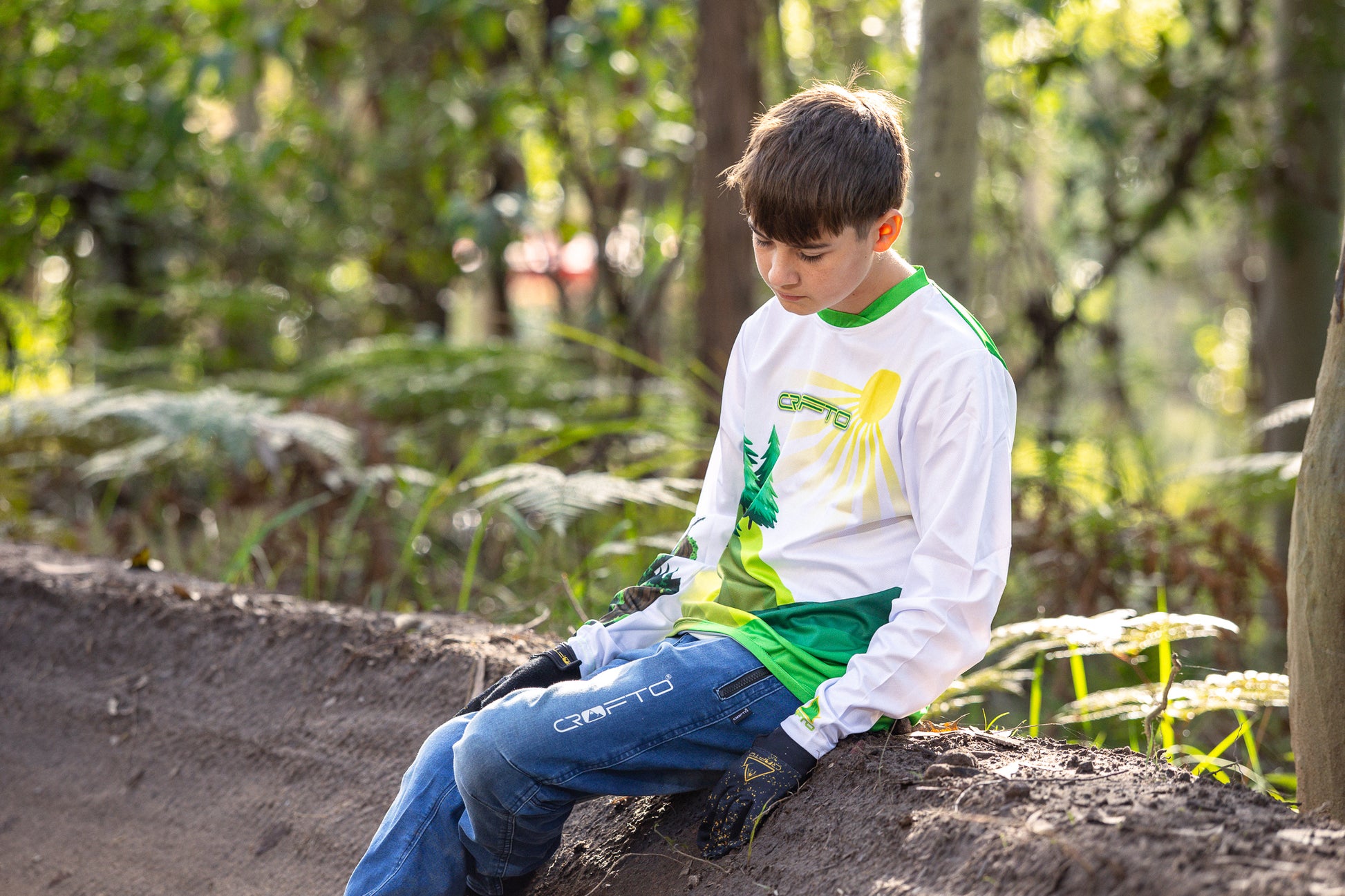 Boy sitting on the edge of a berm in the forest wearing a CROFTO Hacklberg MTB jersey 