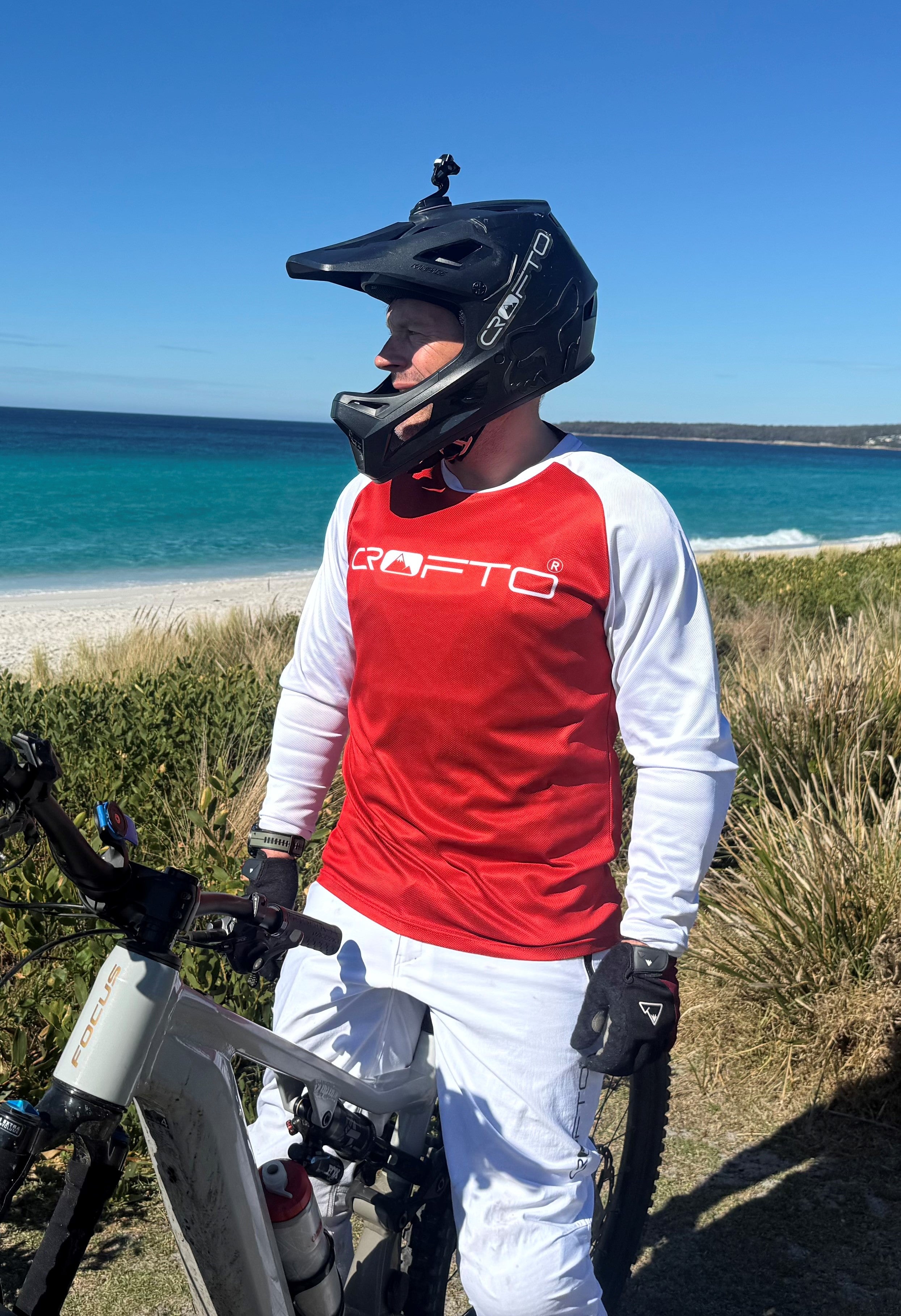 Person wearing a red and white jersey with a helmet, standing next to a bicycle on a beach.