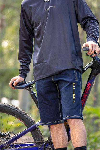 A person wearing a CROFTO Enduro MTB Jersey and CROFTO Denim MTB Jorts shorts, standing next to a mountain bike in an outdoor setting with blurred green trees in the background.