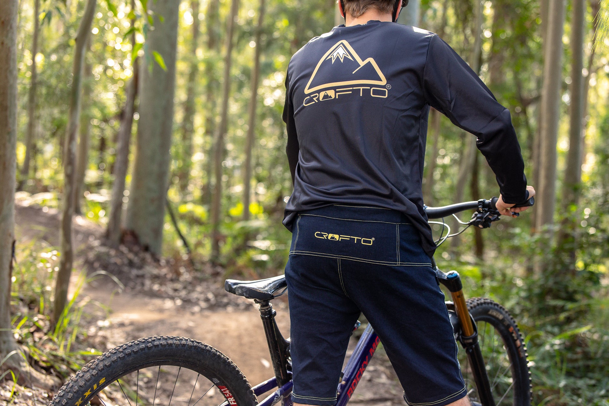 A rear view of a person wearing a CROFTO Enduro MTB Jersey and CROFTO Denim MTB Jorts, Sitting on a mountain bike in the  Australian forest 