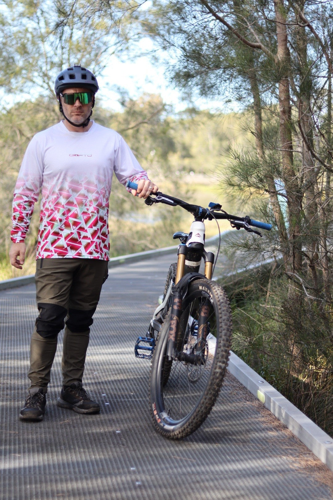 A man standing next to a mountain bike next to the forest wearing a CROFTO Mont Blanc Long Sleeve MTB jersey and dark green Amaroo pants
