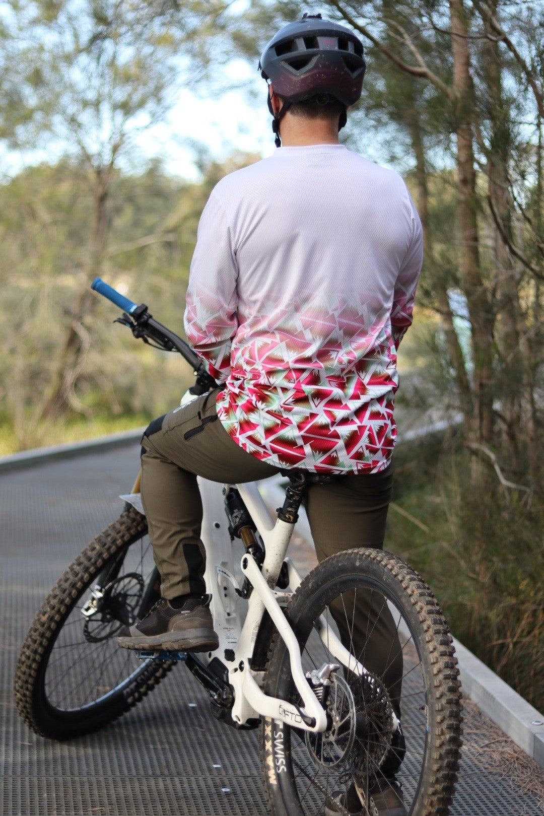 A man sitting on a mountain bike next to the forest  facing away from the camera wearing a CROFTO Mont Blanc Long Sleeve MTB jersey and dark green Amaroo pants