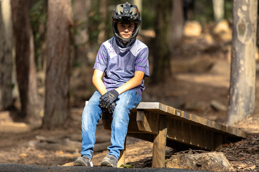 A person wearing CROFTO Slate riding jersey and Crofto MTB Jeans sitting on the edge of a jump in the forest