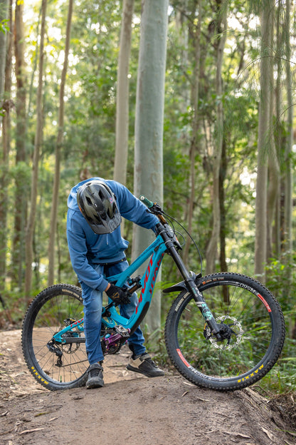 A mountain biker in a blue hoodie and helmet leans over a turquoise full-suspension bike on a dirt trail in a forest.