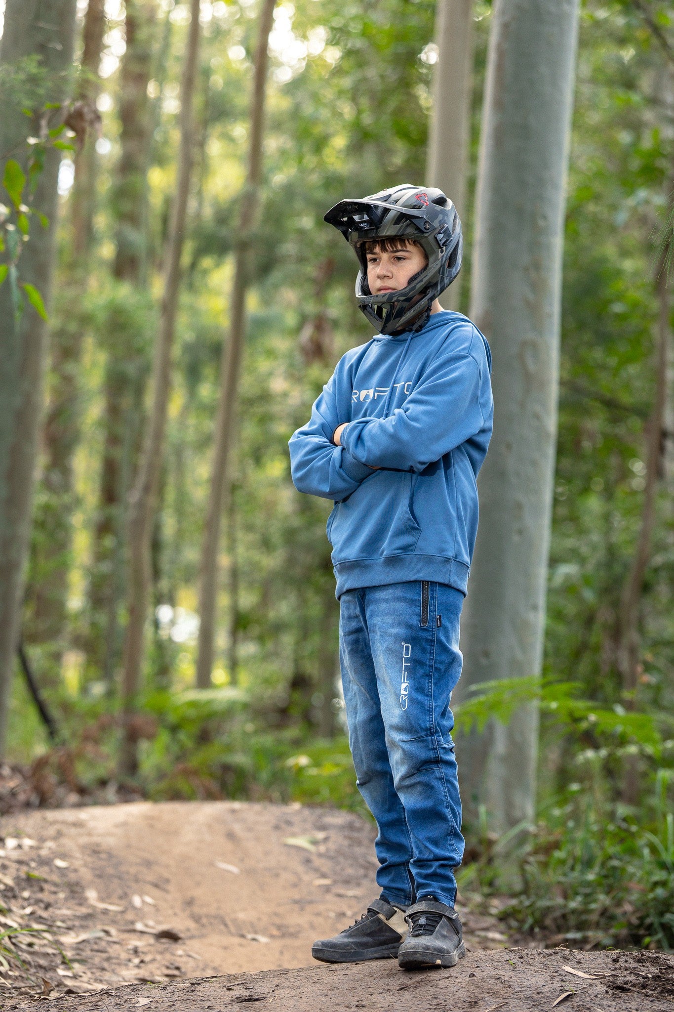 A person standing on a dirt trail in a forest, wearing a black full-face helmet, a blue CROFTO hoodie, blue jeans, and dark sneakers, with arms crossed.