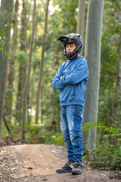 A person standing on a dirt trail in a forest, wearing a black full-face helmet, a blue CROFTO hoodie, blue jeans, and dark sneakers, with arms crossed.