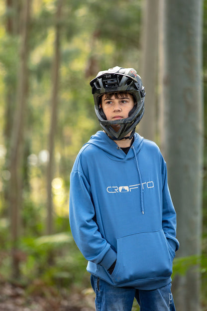 A young man wearing a blue Crofto hoodie and a black full-face helmet stands in a forest setting.