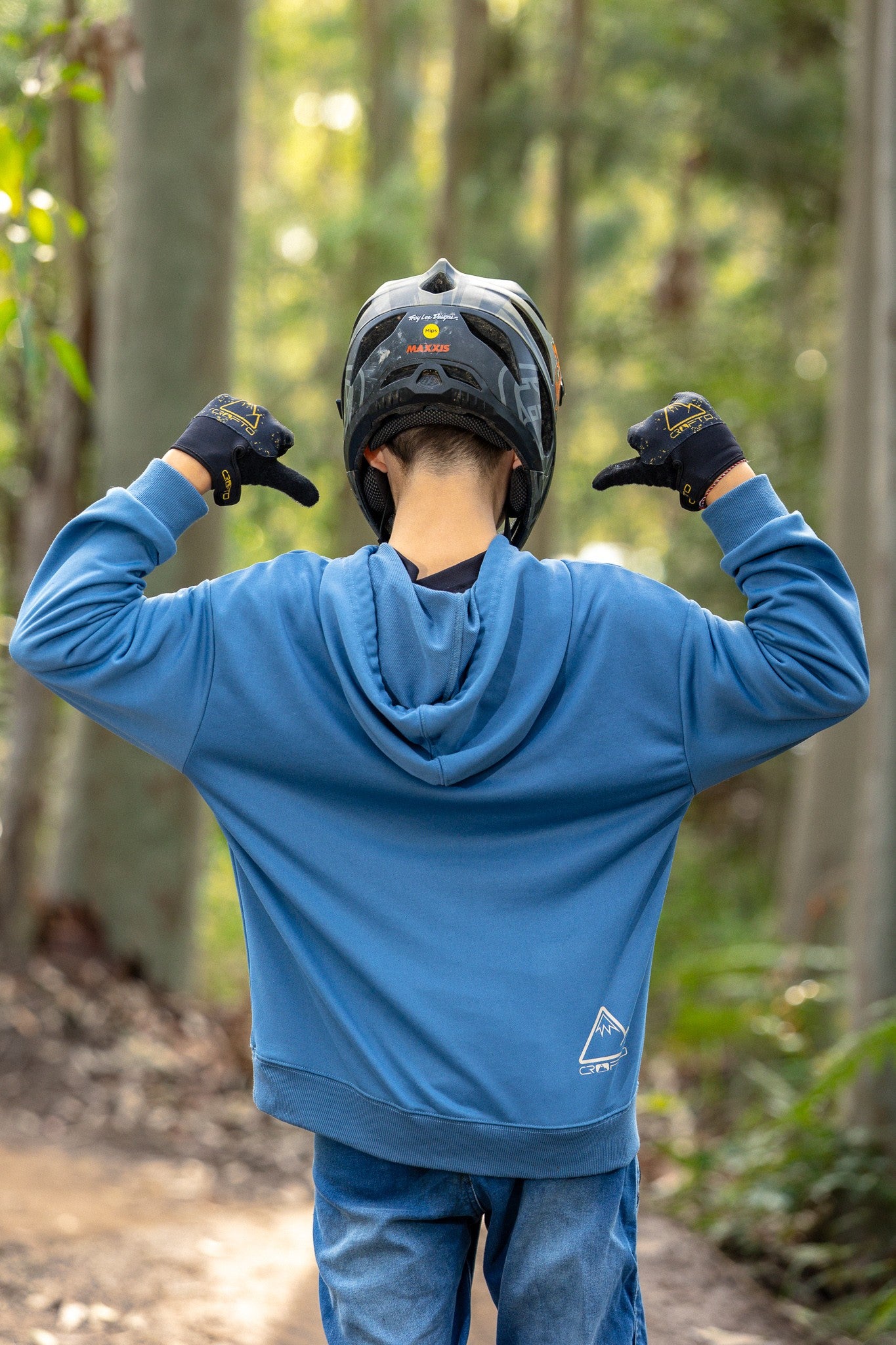 A person viewed from behind wearing a blue hooded sweatshirt, blue jeans, a black helmet, and black gloves, standing on a forest path with hands raised toward the helmet.