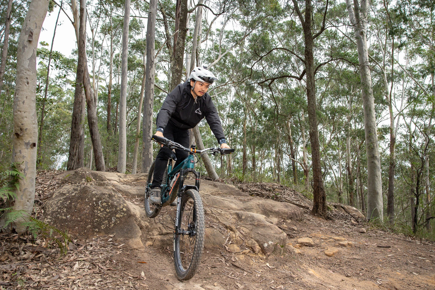A mountain biker wearing a Black crofto trail jacket rides a teal and black bike over a rocky dirt trail in a dense forest.