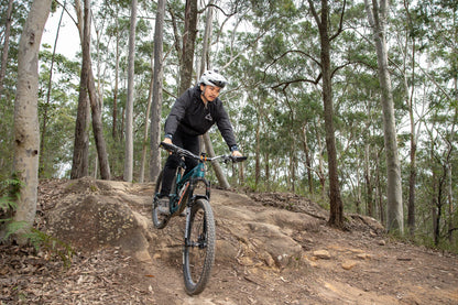 A mountain biker wearing a Black crofto trail jacket rides a teal and black bike over a rocky dirt trail in a dense forest.