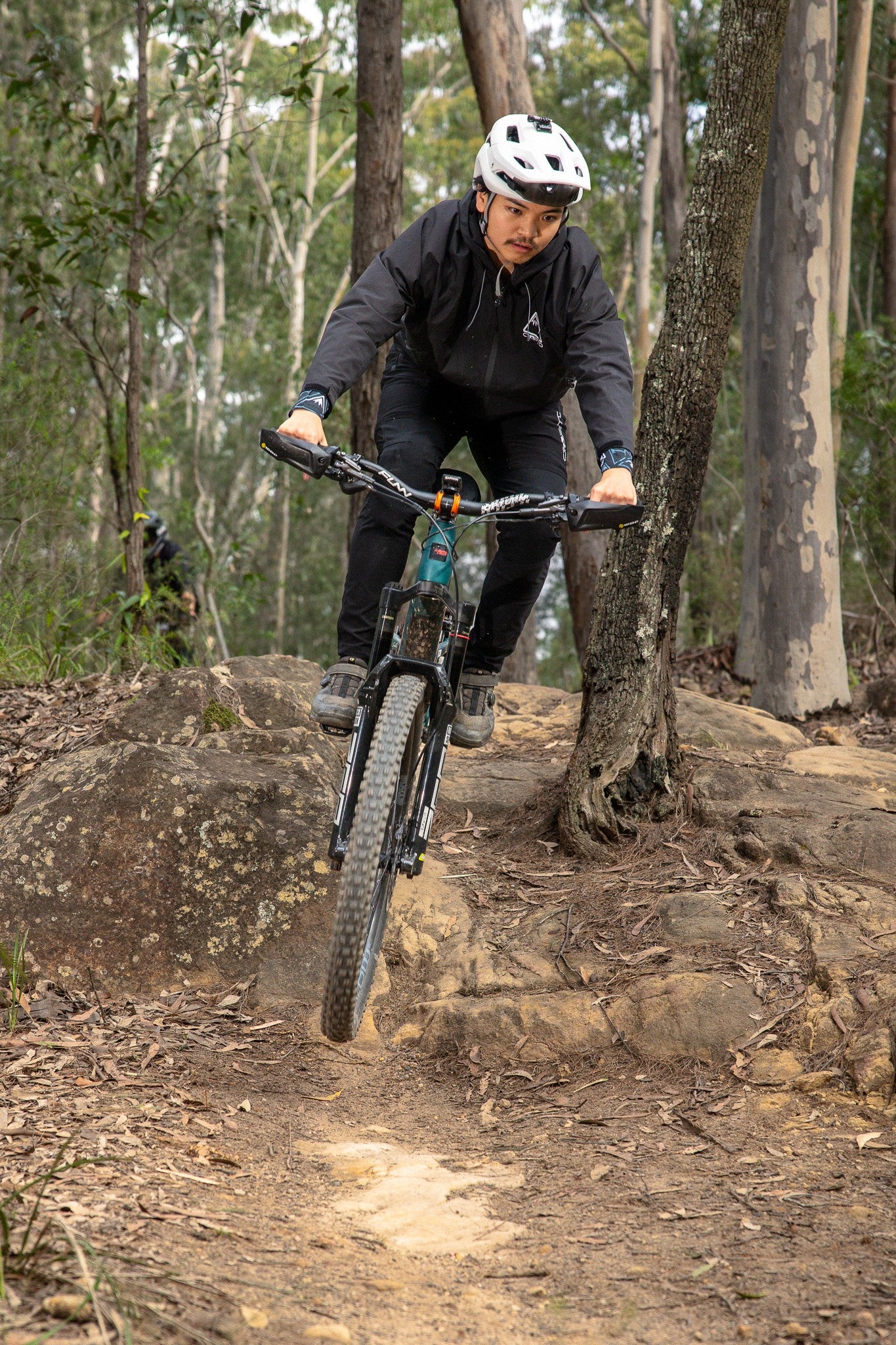 A mountain biker wearing a Black crofto trail jacket rides a teal and black bike over a rocky dirt trail in a dense forest.