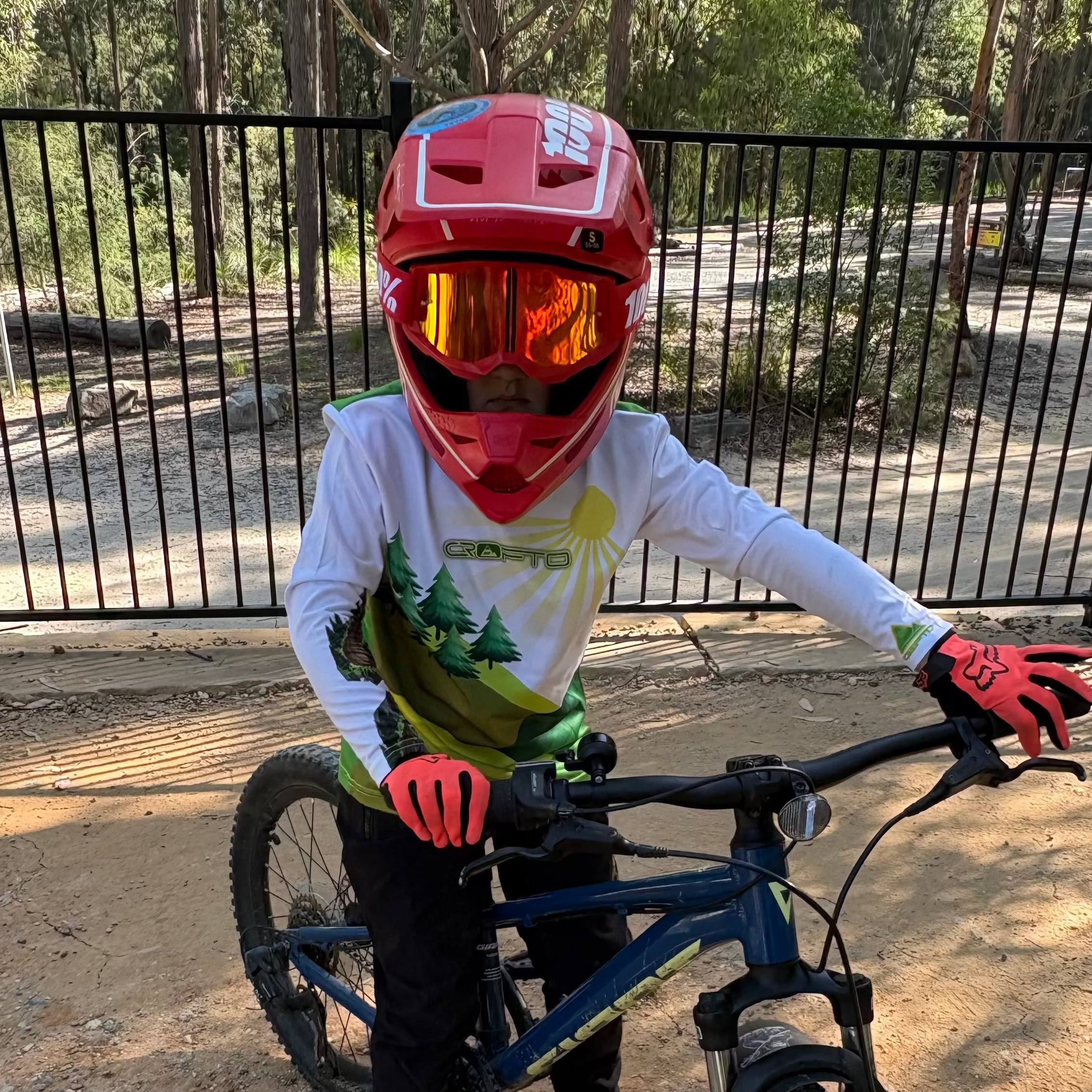 Young mountain biker wearing a CROFTO MTB jersey, red full-face helmet and riding gloves while standing with a mountain bike on a dirt trail in a forest setting.