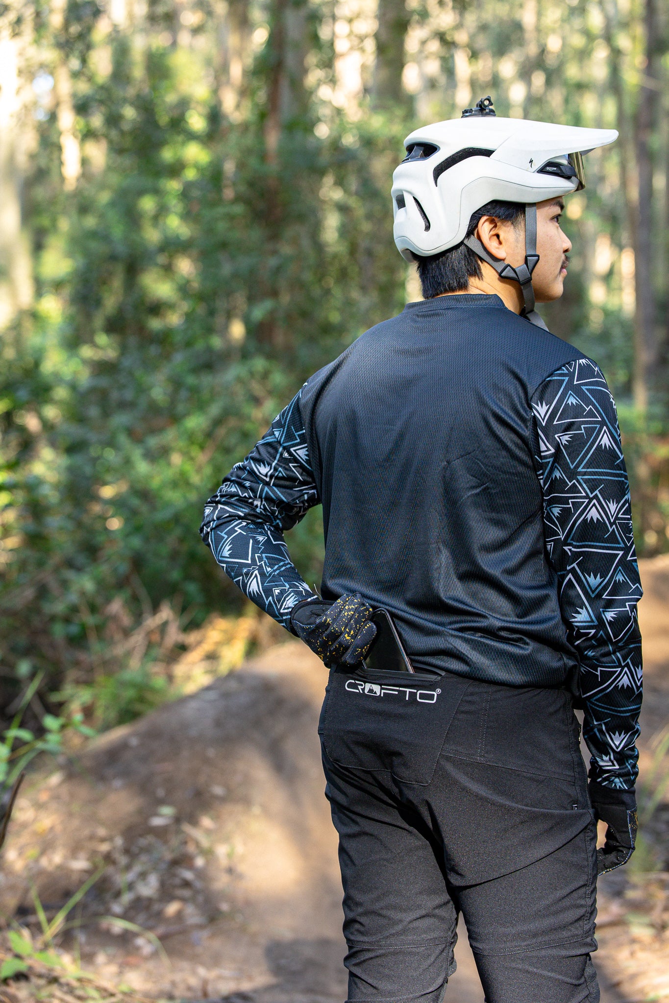A person outdoors wearing a black long-sleeve crofto mozaic jersey and black amaroo pants putting a phone in the rear pocket