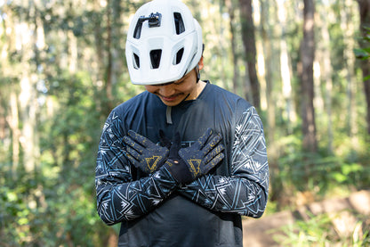 A person wearing a white helmet and a dark long-sleeve Crofto Mozaic jersey  standing in a forest while adjusting Crofto apex pro gloves