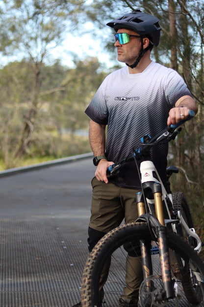 A man wearing a black mountain bike helmet, mirrored sunglasses, and a black-to-white gradient CROFTO Carbon Classic MTB jersey stands next to a mountain bike on by the bush