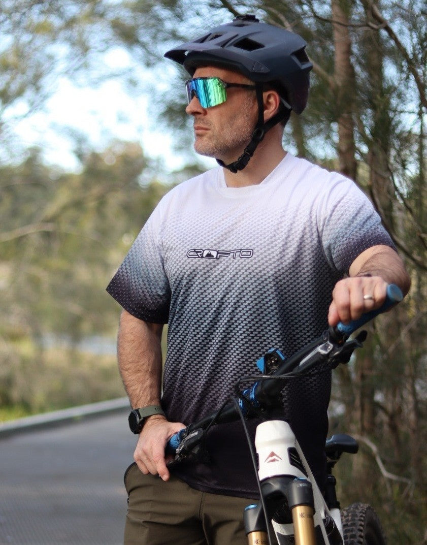 Close up of a man wearing a black mountain bike helmet, mirrored sunglasses, and a black-to-white gradient CROFTO Carbon Classic MTB jersey stands next to a mountain bike on by the bush